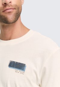 Lichte crème katoenen t-shirt met een grafisch ontwerp in blauw en grijs met de tekst "TOM TAILOR ESTD 1962," ronde halslijn en korte mouwen.