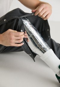 Szürke szövetborítás, alatta látható egy fém protézis láb. Gombok és patentok rögzítik a borítást; egy fehér zokni részben látható.