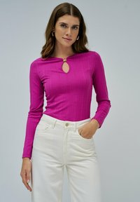 Top de manga larga ajustado en un tejido de canalé rosa vibrante, con un corte en forma de llave y un acento dorado en el escote. Combinado con pantalones blancos de cintura alta.