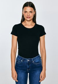 Vrouw met lang bruin haar, gouden oorringen in de vorm van hoepels, een nauwsluitend zwart T-shirt met korte mouwen en een blauwe spijkerbroek, staand tegen een effen achtergrond.