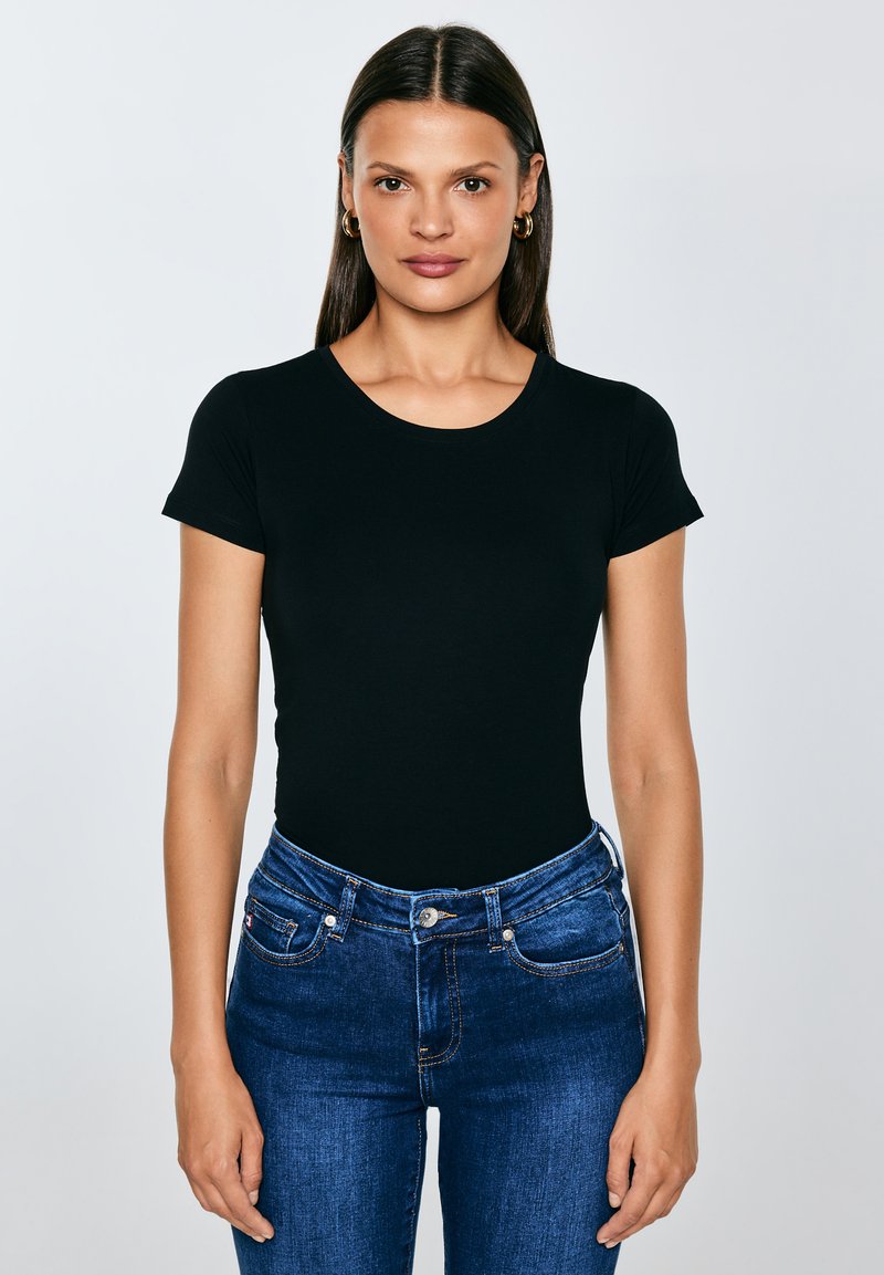 Vrouw met lang bruin haar, gouden oorringen in de vorm van hoepels, een nauwsluitend zwart T-shirt met korte mouwen en een blauwe spijkerbroek, staand tegen een effen achtergrond.