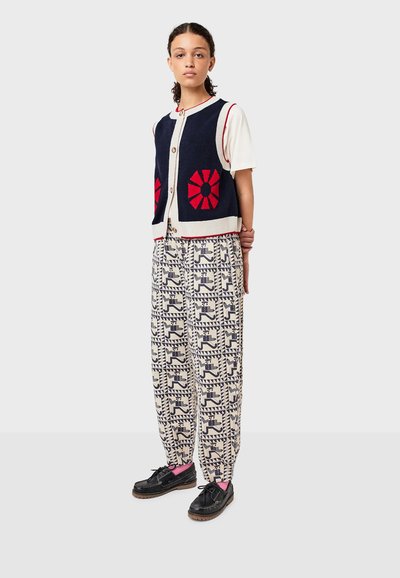Donk blauw gebreid vest met een rood bloemenpatroon over een wit t-shirt, gecombineerd met gestreepte crèmekleurige broek en zwarte schoenen met roze sokken.