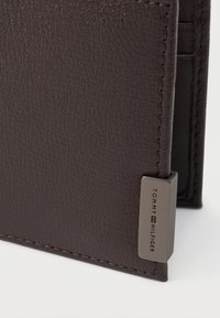 Tmavo hnedá štruktúrovaná kožená peňaženka s rohom a kovovou značkou s vyrytým menom "Tommy Hilfiger" na bielom pozadí.