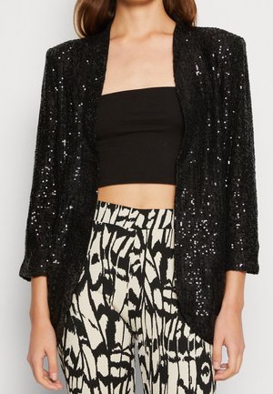 Kobieta w czarnej cekinowej marynarce, czarnym topie bandeau i wysokich spodniach z abstrakcyjnym czarno-białym wzorem.