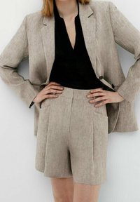 Donna che indossa un blazer di lino beige e pantaloni corti a vita alta coordinati, con una maglietta nera a scollo a V, mani sui fianchi su uno sfondo bianco.