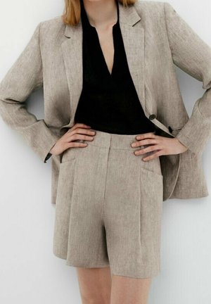 Donna che indossa un blazer di lino beige e pantaloni corti a vita alta coordinati, con una maglietta nera a scollo a V, mani sui fianchi su uno sfondo bianco.