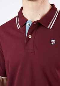Polo shirt bordeaux avec un col bleu clair et des accents rayés. Doté de trois boutons et d'un logo écusson brodé sur la poitrine.