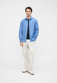 Μπλε ριγωτό πουκάμισο με κουμπιά πάνω από μπλε πουκάμισο, συνδυασμένο με κρεμ παντελόνι και λευκά αθλητικά παπούτσια. Φέρει ένα μικρό κίτρινο λογότυπο στο πουκάμισο.