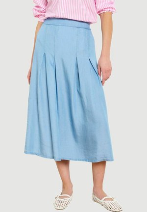 Frau trägt einen knielangen hellblauen plissierten Rock, ein pink-weiß gestreiftes Hemd und weiße geflochtene flache Schuhe vor neutralem Hintergrund.