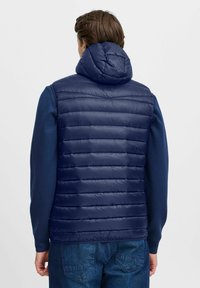 Tumši zils pūkains veste ar noņemamu kapuci, ar kvadrātu tekstūru un elegantu, vieglu dizainu. Aizmugures skats parāda horizontālu šuvju rakstu.