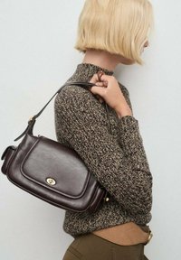 Persona bionda che indossa un maglione di lana marrone e pantaloni marroni, mentre tiene una borsa a tracolla in pelle marrone scuro con chiusura dorata.