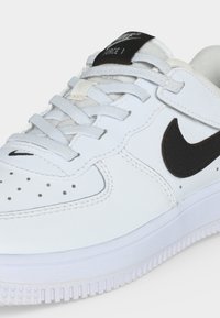 Άσπρο sneaker Nike Air Force 1 με μαύρο λογότυπο Swoosh, λευκά κορδόνια, διάτρητη μύτη και μαύρη ετικέτα στη γλώσσα.