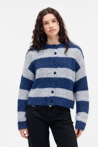 Jonge vrouw met krullend haar, gekleed in een blauw en grijs gestreepte knooptrui en zwarte broek tegen een effen witte achtergrond.