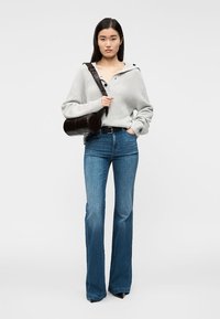 Grå stickad tröja med huva, utsvängda blå jeans i denim och en rund brun handväska. Modellen bär spetsiga svarta klackar. Enkel bakgrund.