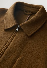 Braune Wolljacke mit Reißverschluss, weitem Kragen, sichtbarem Reißverschlussgriff und einer Stoffschlaufe im Kragen.