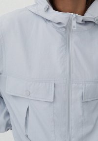 Ljusgrå vindjacka med dragkedja framtill, justerbar huva och två bröstfickor med tryckknappar. Slät, lätt tyg.