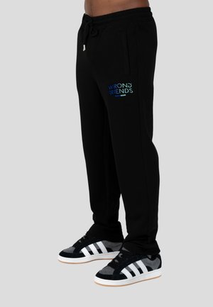 Pantalones de chándal negros con cintura ajustable, presentando el logo "WRONG FRIENDS" en azul y verde en el muslo izquierdo. Combinados con zapatillas negras y blancas.