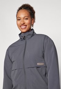 Giacca grigia con colletto alto e zip frontale. Presenta una cucitura orizzontale e una toppa con logo sul lato sinistro. Tessuto liscio e leggero.