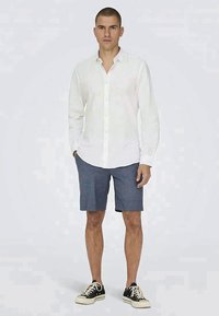 Witte blouse met lange mouwen, gecombineerd met donkerblauwe shorts en zwarte veterschoenen. Glad materiaal met een getailleerde pasvorm.