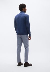 Μπλε πουλόβερ με πλέξη κα λώδιο, ψηλός γιακάς, ανάγλυφο σχέδιο· ανοιχτό γκρι παντελόνι, κανονική γραμμή· σκούρα καφέ παπούτσια με χοντρές σόλες.