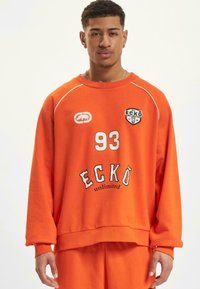 Orange oversized sweatshirt with ribbed cuffs and hem, featuring white accents, logo patches, and text "93 ECKÖ unlimited" on the front.