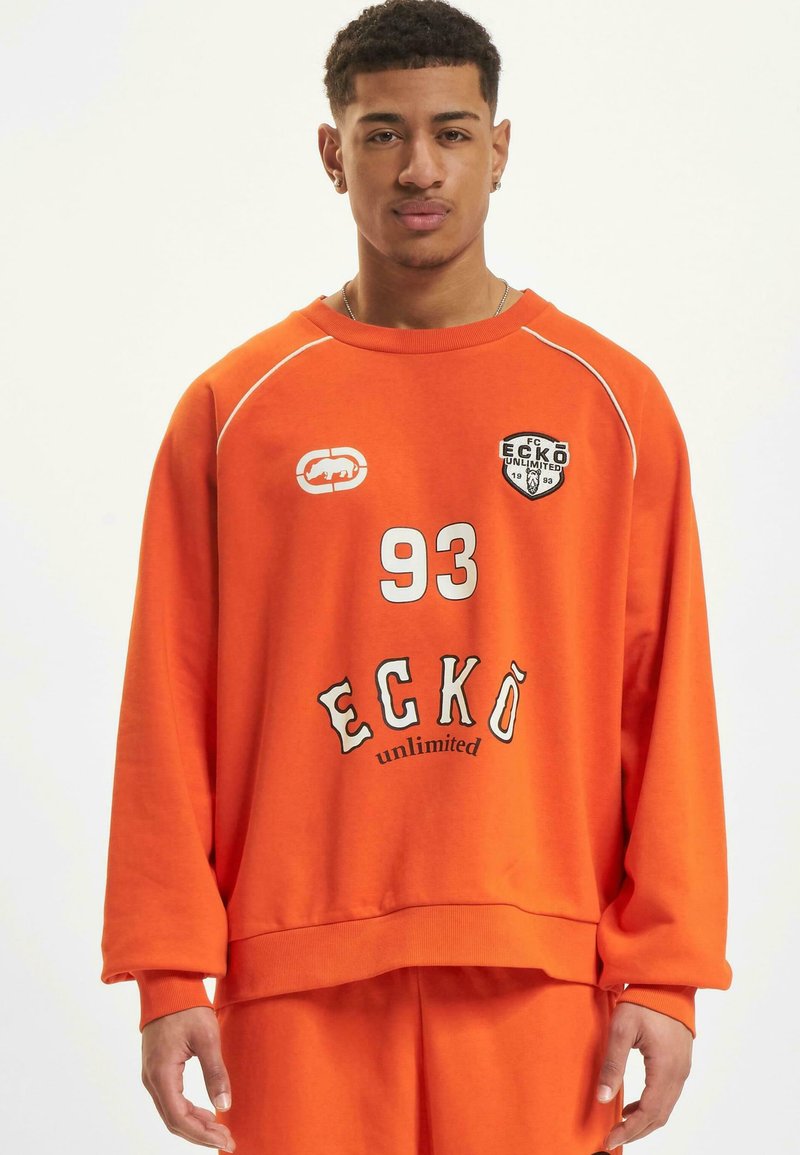 Orange oversized sweatshirt with ribbed cuffs and hem, featuring white accents, logo patches, and text "93 ECKÖ unlimited" on the front.