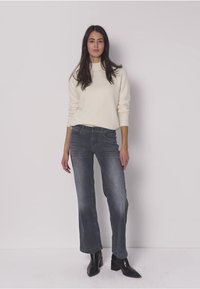 Hellbeiger Pullover kombiniert mit ausgestellten, ausgewaschenen grauen Jeans. Das Modell trägt schwarze Stiefeletten und präsentiert ein lässiges Outfit. Schlichtes, sauberes Design.