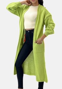 Knälång, öppen cardigan i ljusgrönt stickat material med långa ärmar och två framfickor. Bärs över en vit ribbad topp och mörka byxor.