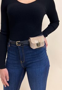 Trblietavý zlatý ledvinka s sponou, připevněná na černém pásku, nošena v pase přes tmavě modré džíny a černý žebrovaný top s dlouhým rukávem.