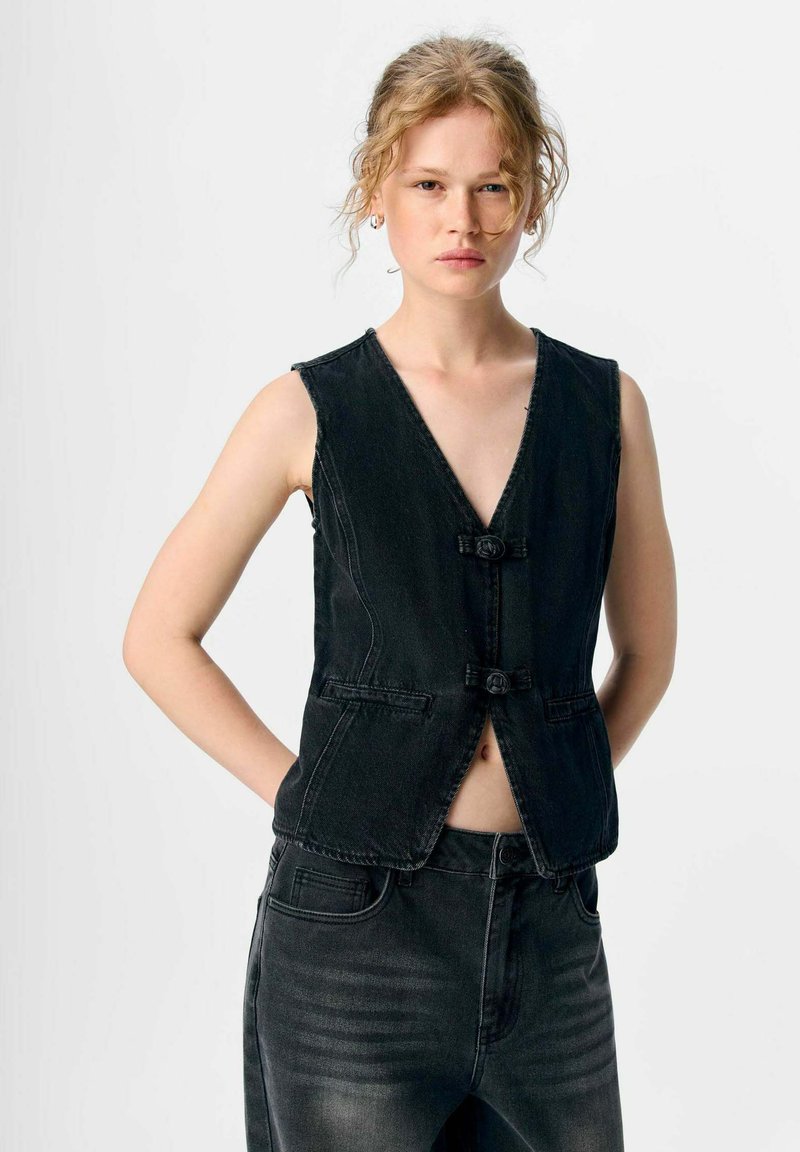 Schwarze Jeansweste mit V-Ausschnitt, ärmellos, mit zwei dekorativen Knöpfen und Taschen; kombiniert mit passenden schwarzen Jeans.