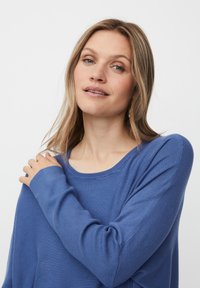 Frau mit hellem Haar, die sanft ihre Schulter berührt, trägt ein blaues Langarmoberteil vor einem schlichten weißen Hintergrund.