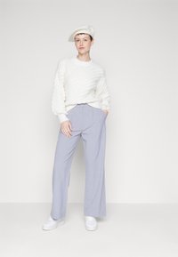 Pullover bianco a trama con un motivo diagonale, pantaloni larghi grigi chiari e sneakers bianche. Un berretto color crema completa l'outfit.