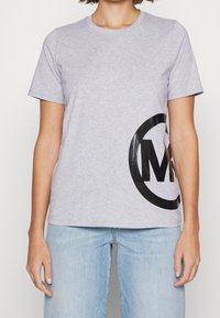 Grijze katoenen t-shirt met korte mouwen, voorzien van een zwart glanzend cirkelvormig logo en de letter "M" op de voorkant. Casual fit, zachte textuur.