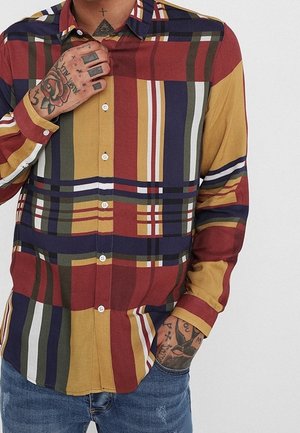 Homme portant une chemise à carreaux multicolore boutonnée avec des tatouages sur les mains et le cou, debout avec une main ajustant le col et l'autre dans la poche.