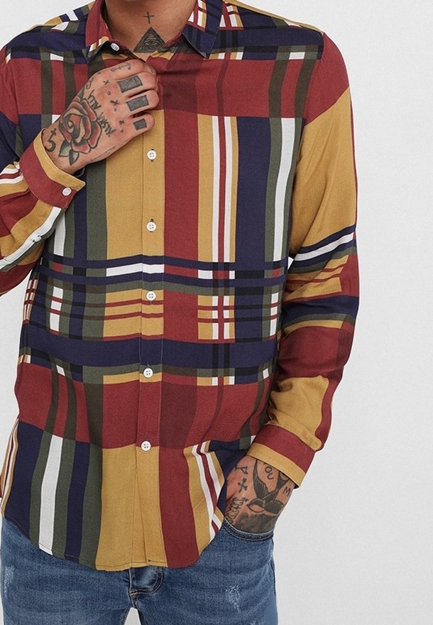 Homme portant une chemise à carreaux multicolore boutonnée avec des tatouages sur les mains et le cou, debout avec une main ajustant le col et l'autre dans la poche.