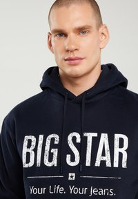 Jaunas vyras trumpais plaukais dėvi tamsiai mėlynę džemperį su gobtuvu, priekyje baltomis raidėmis užrašyta „BIG STAR Your Life. Your Jeans.“