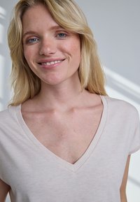 Nasmejana žena plave kose i plavih očiju, odjevena u svijetloružičastu majicu s V-izrezom, na nježno osvijetljenoj neutralnoj pozadini.