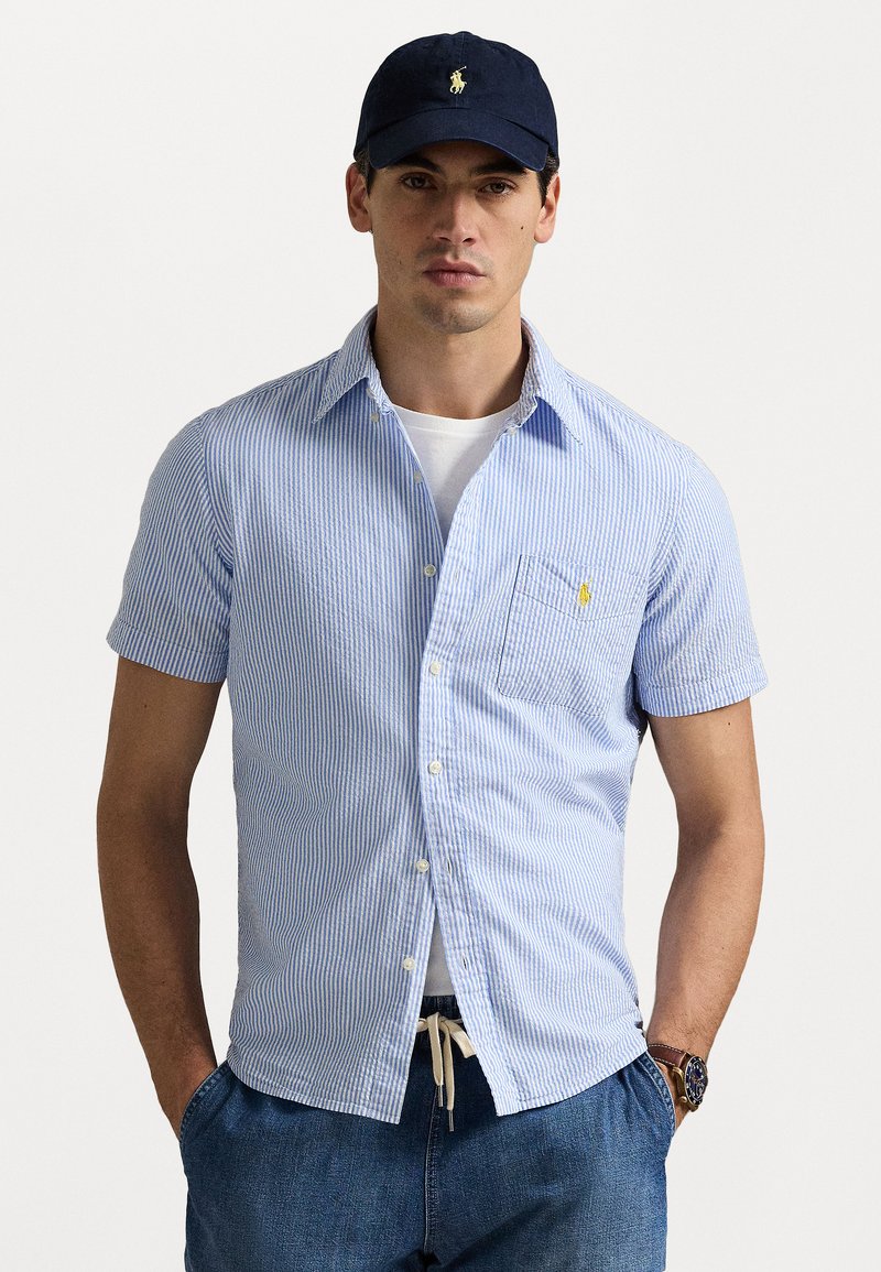 Chemise à manches courtes à rayures bleues et blanches, avec une poche poitrine et un petit logo sur le col. Portée avec un short en denim.