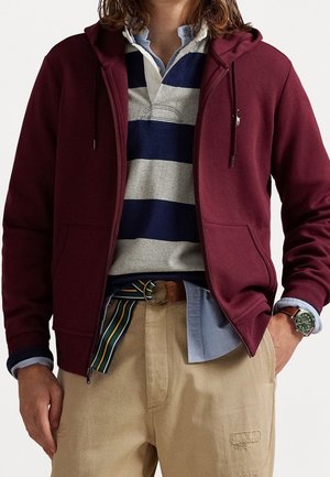Person trägt einen kastanienbraunen Kapuzenpullover mit Reißverschluss, ein blau-weiß gestreiftes Rugbyshirt, beige Hosen mit gestreiftem Gürtel und eine grüne Armbanduhr.