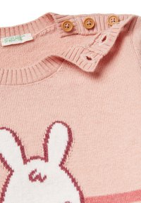 Roze gebreide trui met een wit konijnontwerp, geribde kraag en drie bruine knopen als sluiting bij de schouder. Zachte textuur, casual stijl.