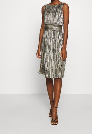 Kvinde iført en ærmeløs, knælængde metalisk plisseret kjole med et bredt bælte og sorte strophæle, stående på et lysegråt gulv.