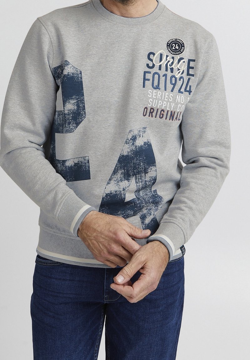 Homme portant un sweat-shirt gris avec un texte imprimé bleu marine et blanc et un grand numéro 24, associé à un jean bleu foncé, mains jointes devant.