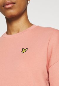 Světle růžová krátkosrstá košile s kulatým výstřihem, na hrudi s malým vyšitým logem ptáčka černé a žluté barvy.