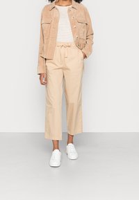 Giacca cropped in suede beige con bottoni e due tasche sul petto, abbinata a pantaloni beige a gamba larga e sneakers bianche. Top a righe visibile.