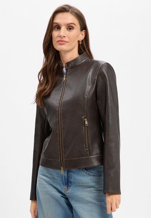 Femme aux longs cheveux bruns portant une veste en cuir marron foncé zippée et un jean bleu, debout devant un fond blanc uni.