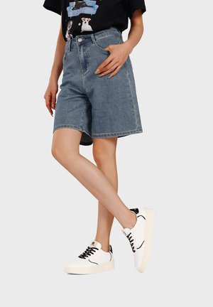 Persona che indossa pantaloncini di jeans di media lunghezza, una maglietta nera con immagini di cani e sneakers bianche con dettagli neri, in piedi su una gamba sola.