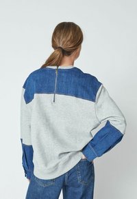 Gray sweatshirt with blue denim accents on shoulders and sleeves. Features a back zipper and cuffs with denim detail. Casual fit.