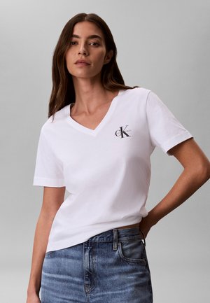 Γυναίκα με μακριά καστανά μαλλιά που φοράει ένα λευκό T-shirt με V λαιμόκοψη Calvin Klein και μπλε τζιν, πόζαρε με το ένα χέρι στη μέση μπροστά σε γκρι φόντο.