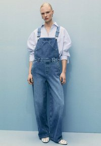 Jeans kombinezon širokog kroja s podesivim naramenicama i džepovima s aplikacijama. Nosite ga preko svijetloplavog prugastog majice i bijelih klompi.