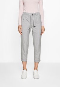 Vrouw in lichtgrijze cropped broek met een stoffen riem, gecombineerd met een lichtroze langemouwen top en witte sneakers.